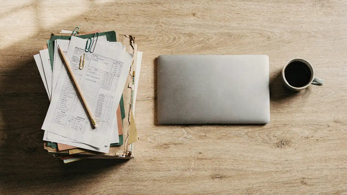 A stack of printed spreadsheets beside a single closed laptop — manual collection replaced by one pipeline
